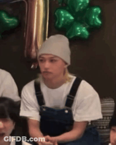 A Man Wearing Overalls And A Beanie Is Sitting In Front Of Green Balloons .. GIF