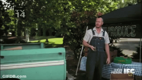 A Man Wearing Overalls Stands In Front Of A Produce Stand GIF