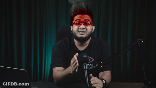 A Man Wearing Red Glasses Is Sitting In Front Of A Microphone With A Foreign Language Behind Him GIF