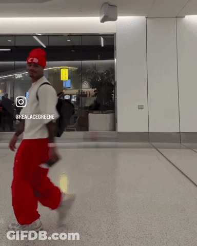 A Man Wearing Red Pants And A Red Hat Is Walking In A Hallway With A Sign That Says @Realacegreene GIF
