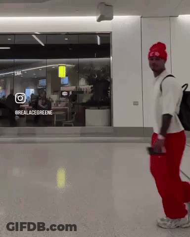 A Man Wearing Red Pants And A White Shirt Is Walking In Front Of A Glass Window That Says @Realacegreene GIF