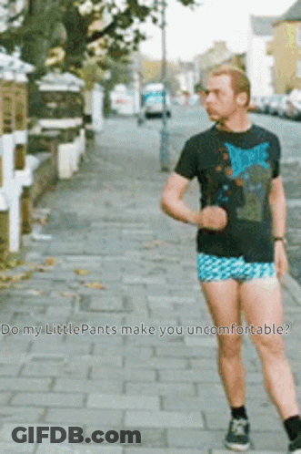 A Man Wearing Shorts And A David Bowie Shirt Is Walking Down A Sidewalk GIF