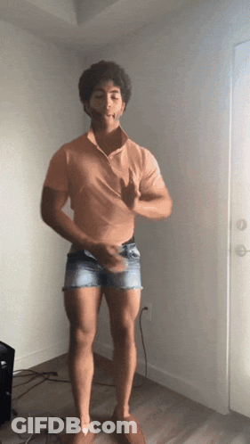 A Man Wearing Shorts And An Orange Shirt Is Standing In Front Of A Door GIF