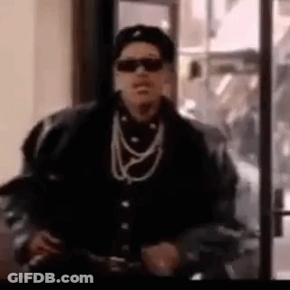A Man Wearing Sunglasses , A Hat , And A Necklace Is Standing In Front Of A Glass Door . GIF