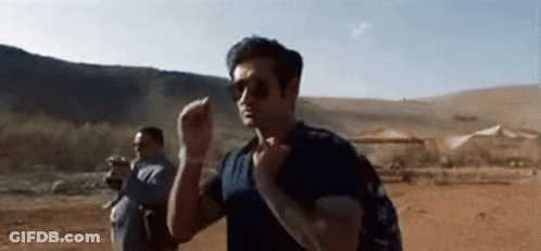 A Man Wearing Sunglasses And A Backpack Is Walking Through A Desert . GIF