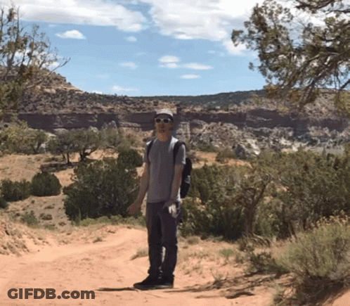 A Man Wearing Sunglasses And A Backpack Stands On A Dirt Road In The Desert GIF