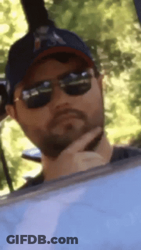 A Man Wearing Sunglasses And A Baseball Cap Is Looking Out Of A Window GIF