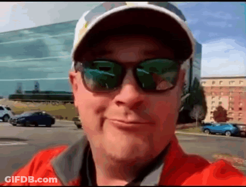 A Man Wearing Sunglasses And A Baseball Cap Is Smiling In Front Of A Building . GIF