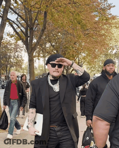A Man Wearing Sunglasses And A Black Beret Is Walking Down A Sidewalk GIF