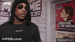 A Man Wearing Sunglasses And A Black Hoodie Is Standing In Front Of A Wall . GIF
