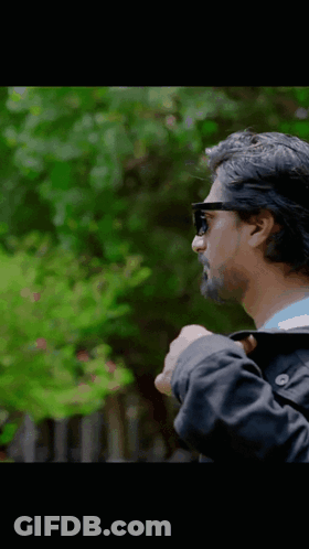 A Man Wearing Sunglasses And A Black Jacket Stands In Front Of Trees GIF