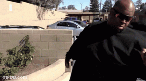 A Man Wearing Sunglasses And A Black Shirt Is Standing In A Parking Lot GIF