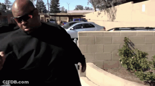 A Man Wearing Sunglasses And A Black Shirt Stands In Front Of A Parking Lot GIF