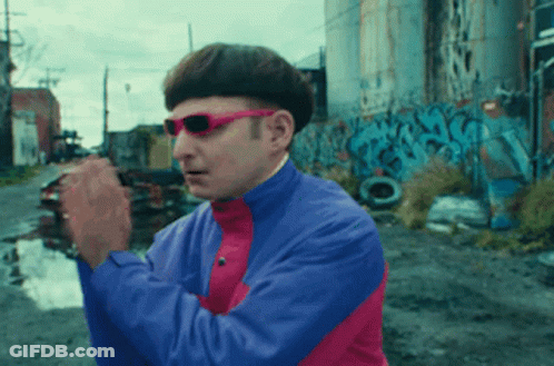 A Man Wearing Sunglasses And A Blue And Pink Jacket Is Standing In Front Of Graffiti GIF