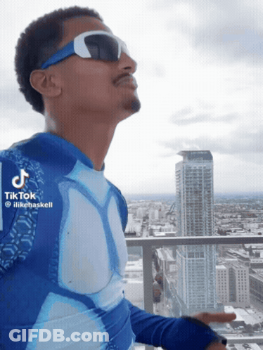 A Man Wearing Sunglasses And A Blue Shirt Is Standing On A Balcony With A City In The Background GIF