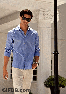 A Man Wearing Sunglasses And A Blue Shirt Is Walking Down A Street GIF