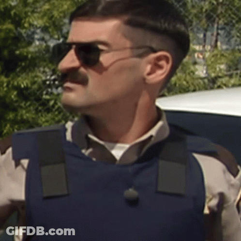 A Man Wearing Sunglasses And A Blue Vest Is Standing In Front Of A Car GIF