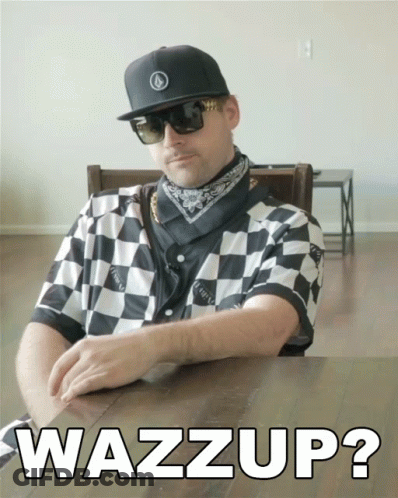 A Man Wearing Sunglasses And A Checkered Shirt Is Sitting At A Table With The Words Wazzup On The Table GIF