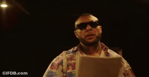 A Man Wearing Sunglasses And A Colorful Shirt Is Standing In A Dark Room Holding A Piece Of Paper . GIF