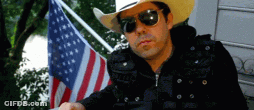 A Man Wearing Sunglasses And A Cowboy Hat Stands In Front Of An American Flag GIF