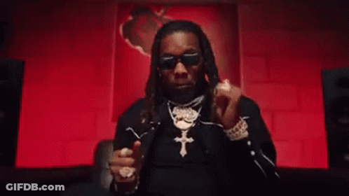 A Man Wearing Sunglasses And A Cross Necklace Is Giving A Thumbs Up In Front Of A Red Wall . GIF