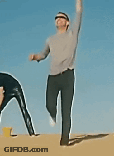 A Man Wearing Sunglasses And A Gray Shirt Is Standing In The Sand GIF