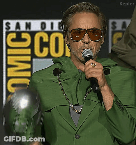 A Man Wearing Sunglasses And A Green Jacket Is Holding A Microphone In Front Of A San Diego Comic Con Sign GIF