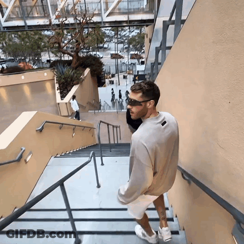 A Man Wearing Sunglasses And A Grey Sweatshirt Is Walking Down Stairs GIF