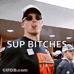 A Man Wearing Sunglasses And A Hat Is Smoking A Cigarette And Says " Sup Bitches " . GIF