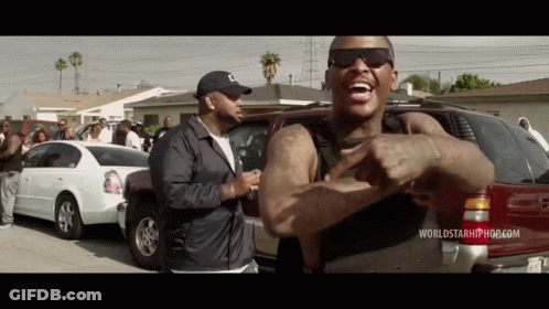 A Man Wearing Sunglasses And A Hat Is Standing In Front Of A Red Suv And A White Car . GIF