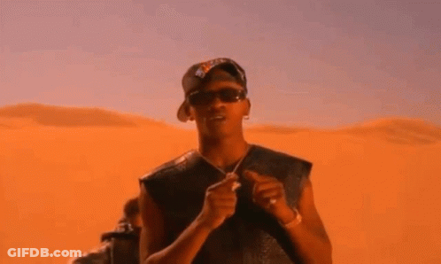 A Man Wearing Sunglasses And A Hat Is Standing In Front Of A Sand Dune . GIF