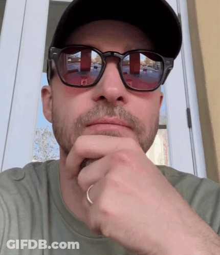 A Man Wearing Sunglasses And A Hat Looks At The Camera With His Hand On His Chin GIF