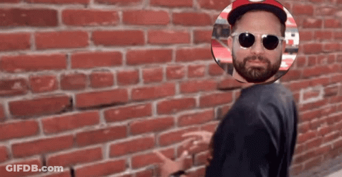 A Man Wearing Sunglasses And A Hat Stands In Front Of A Red Brick Wall GIF