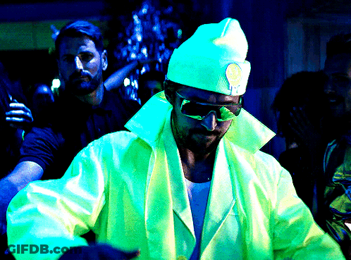 A Man Wearing Sunglasses And A Hat With A Smiley Face On It Stands In A Dark Room GIF