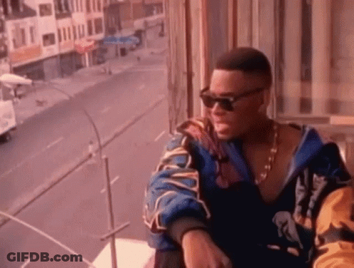 A Man Wearing Sunglasses And A Jacket Is Sitting On A Balcony Overlooking A Street . GIF