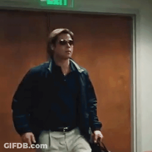 A Man Wearing Sunglasses And A Jacket Is Walking Through A Doorway Holding A Briefcase . GIF