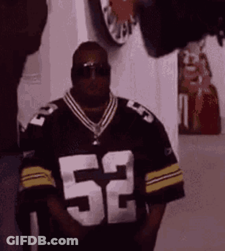 A Man Wearing Sunglasses And A Jersey With The Number 52 On It Is Standing In A Room . GIF