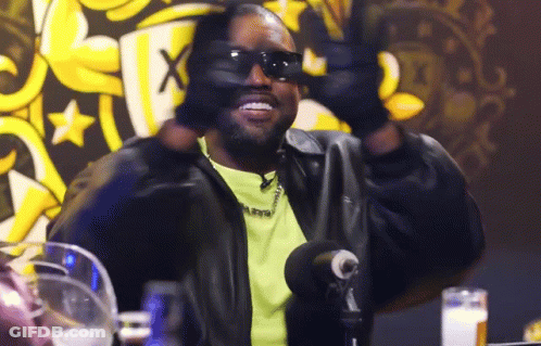 A Man Wearing Sunglasses And A Leather Jacket Is Sitting At A Table With A Microphone . GIF