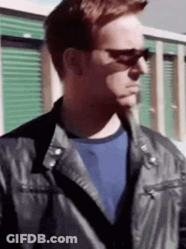 A Man Wearing Sunglasses And A Leather Jacket Stands In Front Of A Green Building . GIF