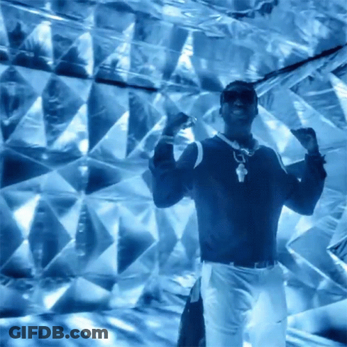 A Man Wearing Sunglasses And A Necklace Is Dancing In Front Of A Blue Wall GIF