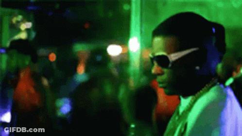 A Man Wearing Sunglasses And A Necklace Is Standing In A Crowded Room . GIF
