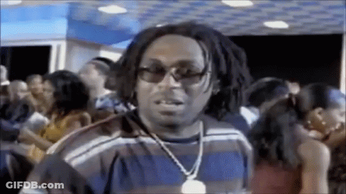 A Man Wearing Sunglasses And A Necklace Is Standing In A Crowded Room . GIF