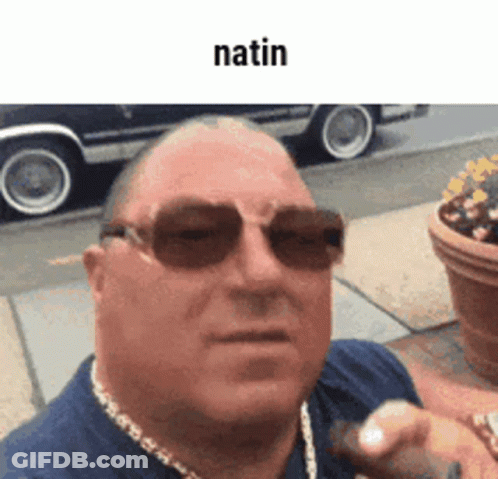 A Man Wearing Sunglasses And A Necklace Is Standing On A Sidewalk In Front Of A Car . GIF