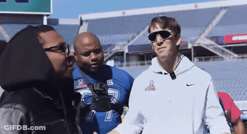 A Man Wearing Sunglasses And A Ny Giants Jersey Talks To Another Man GIF