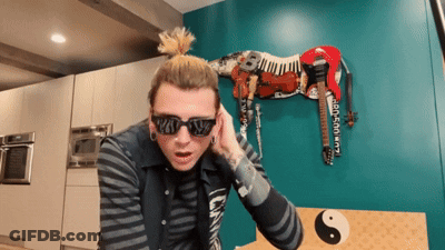 A Man Wearing Sunglasses And A Ponytail Stands In Front Of A Zebra Statue GIF