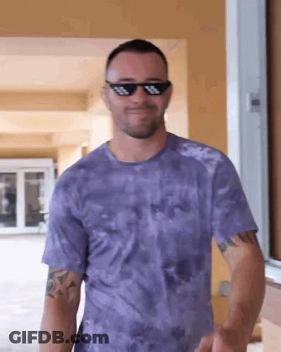 A Man Wearing Sunglasses And A Purple Tie Dye Shirt Is Smiling GIF