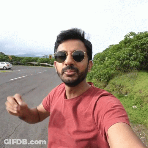 A Man Wearing Sunglasses And A Red Shirt Is Standing On The Side Of A Road GIF