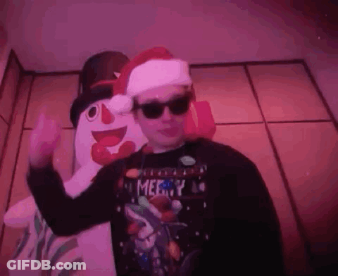 A Man Wearing Sunglasses And A Santa Hat Is Standing In Front Of A Snowman . GIF