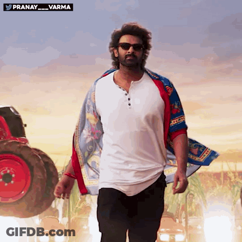 A Man Wearing Sunglasses And A Scarf Is Walking In Front Of A Red Tractor With The Words Pranay Varma Above Him GIF