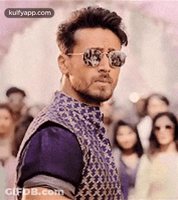 A Man Wearing Sunglasses And A Vest Is Standing In Front Of A Crowd Of People . GIF